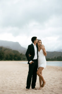 Wedding Photographer, groom in tux and bride in white short dress stand barefoot on the beach sand